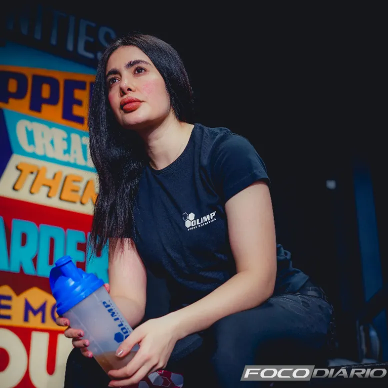 Jovem mulher em roupas de esporte segurando uma garrafa de shake de proteína, com um fundo motivacional de academia.
